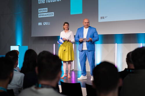 Zwei Moderatoren präsentieren auf einer IFA-2025-Bühne vor Publikum, sie halten Notizen in den Händen und stehen im Schein farbiger LED-Lichtstreifen.