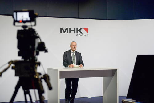 Geschäftsmann im Anzug hält eine Präsentation an einem modernen Rednerpult vor dem Logo der MHK Group, während er von einer Kamera im Studio gefilmt wird