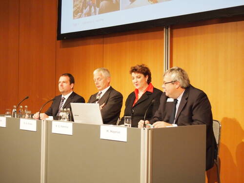 Vier Konferenzteilnehmende sitzen hinter einem Podium, sprechen in Mikrofone und schauen auf einen Laptop, während sie eine Präsentation vor einer holzvertäfelten Wand halten.