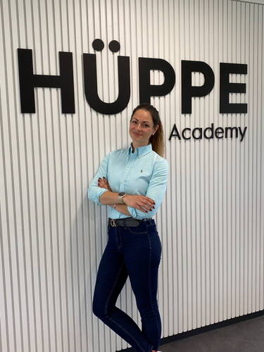 Selbstbewusste Frau mit verschränkten Armen lehnt vor der Wand mit dem Schriftzug „HÜPPE Academy“ in einem modernen Büroflur