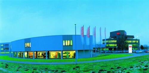 Modernes Firmengebäude mit großen Fenstern und beleuchteten Innenräumen neben einem mehrstöckigen Bürokomplex in einer weitläufigen grünen Landschaft bei Abenddämmerung