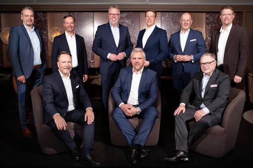 Neun geschäftsmänner in dunklen Anzügen posieren als Managementteam in einem eleganten Konferenzraum, drei sitzend in Sesseln vorne, sechs stehend dahinter vor holzvertäfelter Wand mit Marmorelementen.
