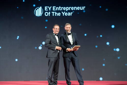 Zwei Männer im Smoking stehen auf rotem Teppich vor der Leinwand mit dem Schriftzug „EY Entrepreneur Of The Year“ und halten eine Auszeichnung in den Händen.