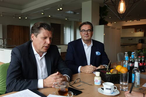 Zwei Geschäftsleute im Anzug sitzen in einem modernen Konferenzraum an einem Tisch mit Getränken und Obstkorb und wirken vertieft in eine Besprechung