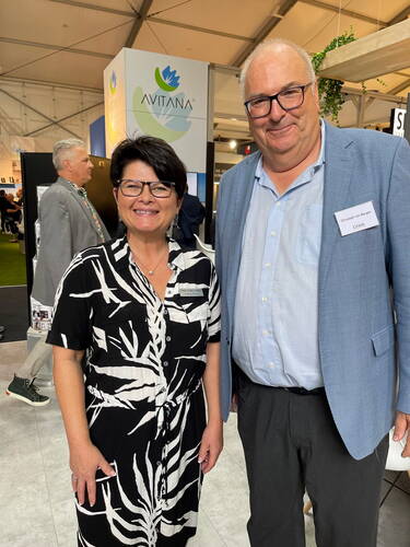Zwei lächelnde Messebesucher stehen nebeneinander vor einem AVITANA‑Messestand in einer hellen Ausstellungshalle.