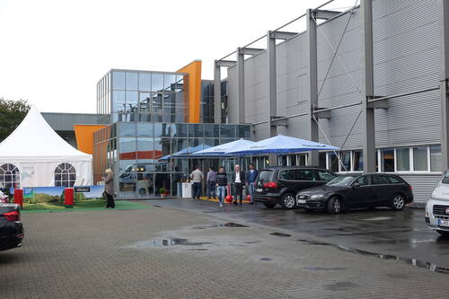 Menschen verlassen nach einem Regenschauer ein modernes Firmengebäude mit Glasfassade, vor dem Autos geparkt sind und blaue Pavillonzelte sowie ein weißes Veranstaltungszelt aufgebaut sind.