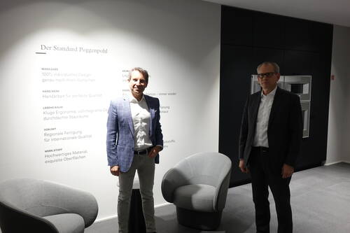 Zwei Geschäftsleute stehen in einem modern gestalteten Showroom von Poggenpohl vor einer beleuchteten Wand mit Unternehmensleitlinien und zwei grauen Designersesseln