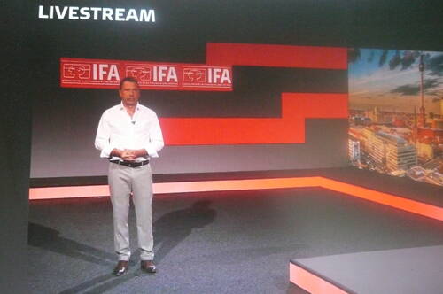 Moderator steht in einem modernen Studio vor IFA-Logos und einer Berlin-Panoramagrafik während eines Livestreams