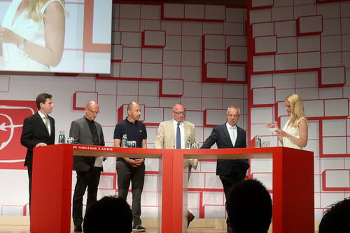 Moderatorin führt eine Podiumsdiskussion mit fünf männlichen Experten an einem roten Stehtisch auf einer Konferenzbühne mit geometrischem Hintergrund.
