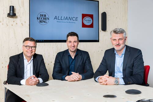 Drei Geschäftsmänner in Anzügen sitzen lächelnd an einem Konferenztisch vor einem Bildschirm mit den Logos von Der Küchen Ring, Alliance und KüchenTreff