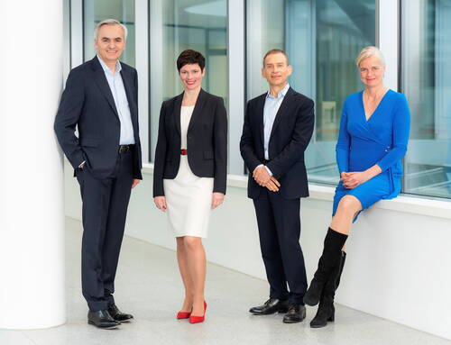 Vier Geschäftsleute in formeller Kleidung posieren lächelnd in einem modernen, hellen Büroflur, drei stehend und eine Frau im blauen Kleid auf einer Fensterbank sitzend.
