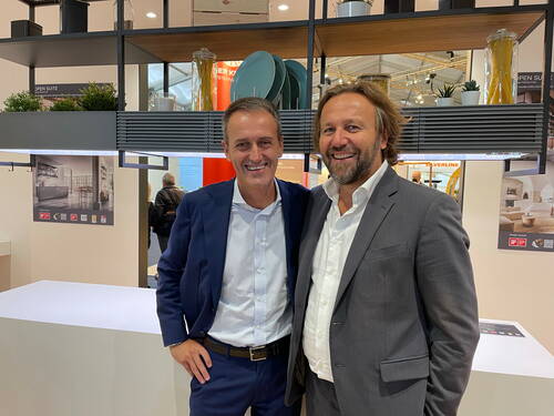 Zwei lächelnde Männer in Anzügen stehen Arm in Arm in einer modernen Messestand-Küche mit Regalsystem, Deko-Pflanzen und Geschirr im Hintergrund