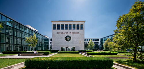 Modernes Verwaltungsgebäude des Flughafens München mit zentralem Uhrenturm, umgeben von symmetrischen Glasfassaden, Grünanlagen und Sitzbereichen bei blauem Himmel