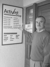 Mann in Pullover steht neben einem Firmenschild der Activent Group und zeigt mit der Hand auf die aufgelisteten Unternehmensbereiche an einer Hauswand.