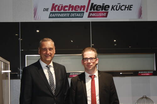 Zwei Geschäftsmänner im Anzug stehen lächelnd in einer modernen Küchenausstellung vor einem „Küchen Keie“-Firmenschild