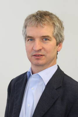 Mittelalterlicher Mann mit kurzen grauen Haaren im dunkelgrauen Sakko und hellblauen Hemd blickt neutral in die Kamera vor hellem Hintergrund