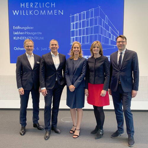 Fünf elegant gekleidete Personen stehen lächelnd nebeneinander vor einer blauen Präsentationswand zur Eröffnungsfeier des Liebherr-Hausgeräte Kundenzentrums.