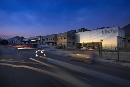 Beleuchteter Industriekomplex mit Elica-Logo an der Fassade bei einbrechender Dämmerung, während Autos Lichtspuren auf der vorbeiführenden Straße hinterlassen