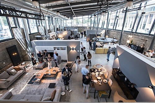 Menschen schlendern durch eine moderne, loftartige Möbel- und Designausstellung mit offenen Wohnbereichen, Sofas und Esstischen unter einer hohen Glasdecke.