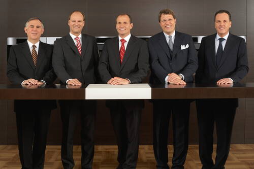 Fünf elegant gekleidete Geschäftsmänner stehen nebeneinander hinter einem modernen Konferenztisch und lächeln formell in die Kamera.
