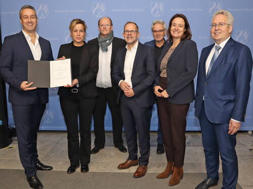 Gruppe von sieben Geschäftsleuten steht vor einer blauen Nord­rhein-Westfalen-Wand, während zwei Personen links gemeinsam eine geöffnete Urkunde in die Kamera halten