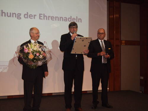 Drei Männer im Anzug stehen auf einer Bühne, einer hält einen Blumenstrauß, der zweite spricht mit Mikrofon und Präsentationsrahmen, während der dritte eine Auszeichnung in der Hand hat.