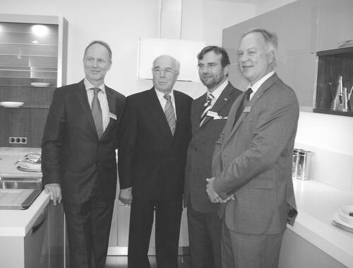 Vier elegant gekleidete Männer im Anzug posieren lächelnd nebeneinander in einer modernen Ausstellungsküche