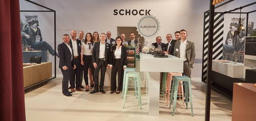 Gruppe von Geschäftsleuten in Anzügen und Businesskleidung posiert lächelnd vor einem Messestand der Marke „Schock“ mit hoher Theke, pastellfarbenen Barhockern und großformatigen Waschbär-Illustrationen im Hintergrund.