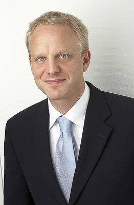 Blonder Mann im dunklen Anzug mit hellblauer Krawatte lächelt leicht vor einer weißen Wand in einem Porträt im Halbprofil.