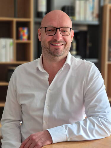 Glatzköpfiger Mann mit Brille und weißem Hemd lächelt entspannt an einem Holztisch in einem modernen Büro mit Bücherregal im Hintergrund