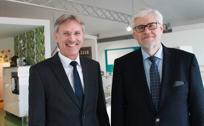 Zwei ältere Männer in Anzügen stehen lächelnd nebeneinander in einem modernen, hellen Büro- oder Ausstellungsraum.