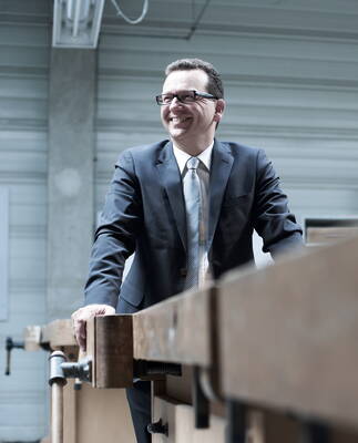 Lächelnder Geschäftsmann im Anzug steht an einer großen Werkbank in einer Industriewerkhalle.