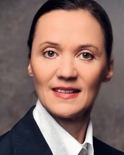 Selbstbewusste, mittelalte Frau mit zurückgebundenen dunklen Haaren und dezentem Make-up im dunklen Blazer und weißer Bluse vor neutralem Hintergrund