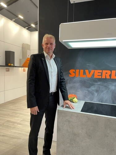 Mann im schwarzen Anzug steht in moderner Ausstellungsküche neben Kochfeld und Dunstabzugshaube mit leuchtendem SILVERLINE-Schriftzug auf der Rückwand