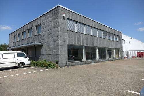 Zweigeschossiges graues Gewerbegebäude mit großen Schaufenstern und Rollläden vor den oberen Fenstern, davor ein leerer Parkplatz mit geparktem weißen Transporter an einem sonnigen Tag.