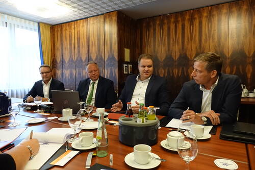Vier Geschäftsleute im Anzug diskutieren angeregt an einem Konferenztisch mit Unterlagen, Laptops und Getränken in einem holzvertäfelten Besprechungsraum.