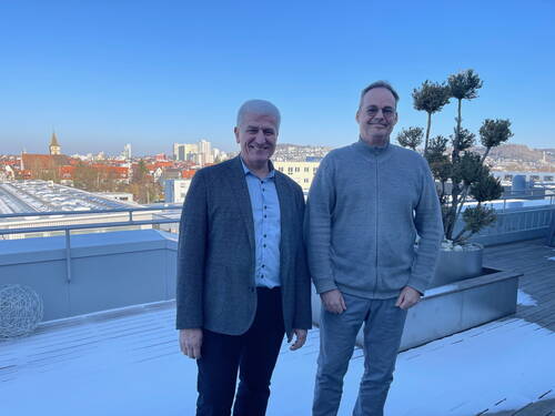 Zwei lächelnde Männer stehen auf einer verschneiten Dachterrasse mit Blick über eine Stadt mit roten Dächern, Kirchturm und klar blauem Himmel.
