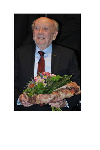 Älterer Mann im Anzug sitzt lächelnd und hält einen großen Blumenstrauß in den Händen