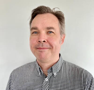 Mittelalterlicher Mann mit kurzen, seitlich gekämmten Haaren lächelt dezent und trägt ein schwarz-weiß kariertes Hemd vor hellem Hintergrund