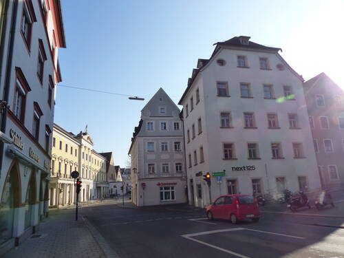 Sonnige Kreuzung einer ruhigen Innenstadtstraße mit historischen mehrstöckigen Häusern, roter Ampel und einem roten Auto im Vordergrund