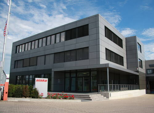 Moderner grauer Bürokomplex mit großflächigen Fenstern, Treppenaufgang und dem Firmenlogo „ninka“ im Eingangsbereich unter blauem Himmel