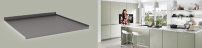 Dunkelgraue, flache Metall-Tablettauflage neben einer modernen, hellen Wohnküche mit mintgrünen Fronten und Kochinsel, an der eine Frau kocht.
