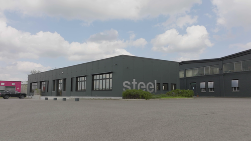Graues Industriegebäude mit der Aufschrift „steel“ an der Fassade und großem, leerem Parkplatz unter bewölktem Himmel