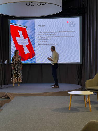 Zwei Personen präsentieren auf einer Messebühne vor einer großen Leinwand mit Schweizer Flagge und Text zur „swiss area“ der Swiss Interior Expo.