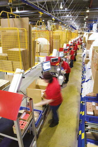 Mitarbeiter in roter Arbeitskleidung verpacken Waren an einer langen Förderstrecke in einer hell beleuchteten Logistikhalle mit gelben Sicherheitsgittern und Computerarbeitsplätzen