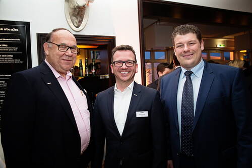 Drei geschäftlich gekleidete Männer lächeln bei einer Abendveranstaltung in einem Restaurant in die Kamera, im Hintergrund sind weitere Gäste und eine Bar zu sehen.