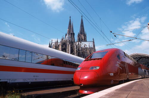 Roter Thalys-Hochgeschwindigkeitszug und weißer ICE stehen nebeneinander im Bahnhof, im Hintergrund ragen die Türme des Kölner Doms in den blauen Himmel.