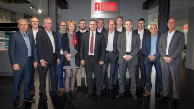 Gruppe von Geschäftsleuten in Anzügen und Businesskleidung posiert lächelnd für ein Gruppenfoto in einem modernen Firmenshowroom mit leuchtendem ALNO-Logo im Hintergrund