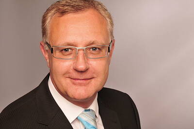 Mittelalterlicher Mann mit Brille im dunklen Anzug und hellblauer Krawatte lächelt freundlich vor neutralem Hintergrund