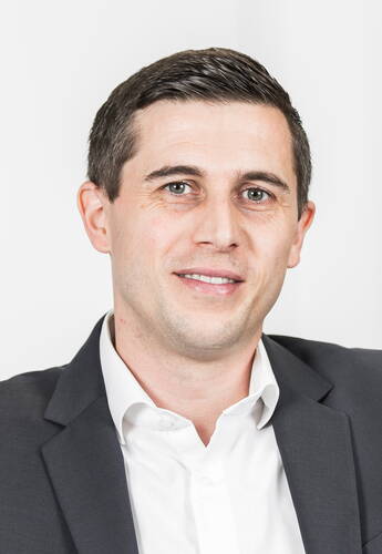 Junger Mann im dunklen Anzug und weißem Hemd lächelt freundlich in einem hellen, neutralen Studiohintergrund